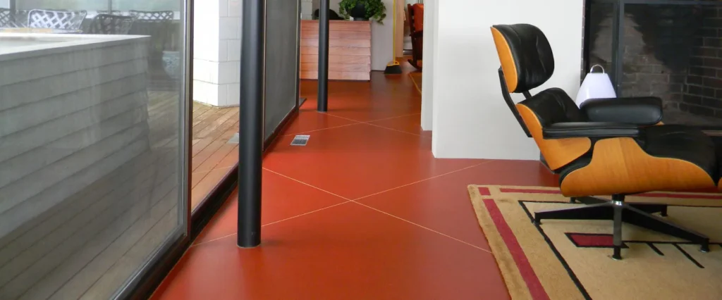 Open-plan Dallas-area space with terracotta red scored concrete overlay floor, Eames lounge chair, brick fireplace, and glass sliding doors — Duraamen decorative concrete flooring